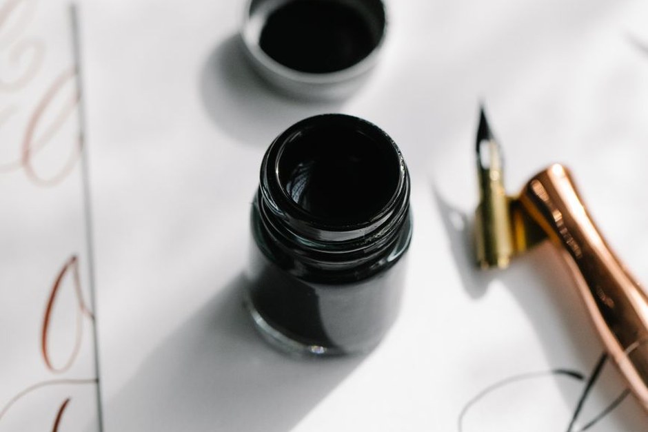 Open bottle of black ink beside a gold fountain pen nib on white paper