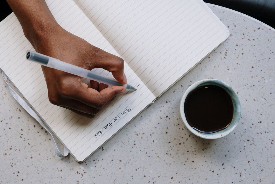 Hand writing a plan in an open notebook beside a cup of coffee