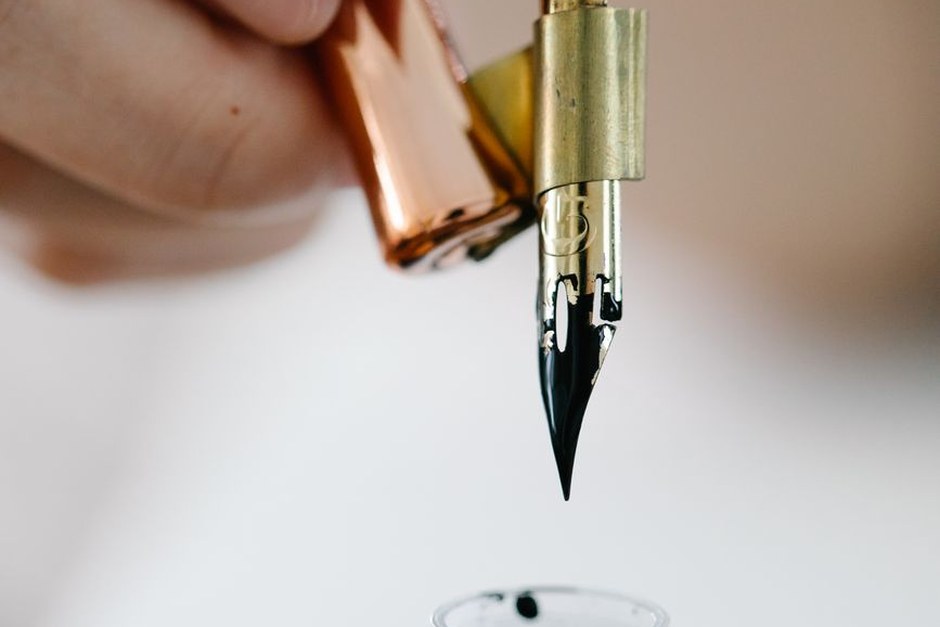 Dip pen nib with a drop of dark ink held above a page